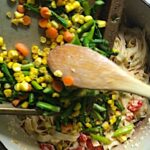 Wood spoon scraping asparagus, corn, and carrots from a sheet pan into the pot of Creamy Pasta Primavera. It has fettuccine, parmesan cheese, roasted asparagus, carrots, tomatoes and fresh corn kernels.