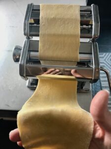 Thick piece of pasta dough going through the pasta roller with hand catching the dough at the bottom for Easy Homemade Pasta recipe.