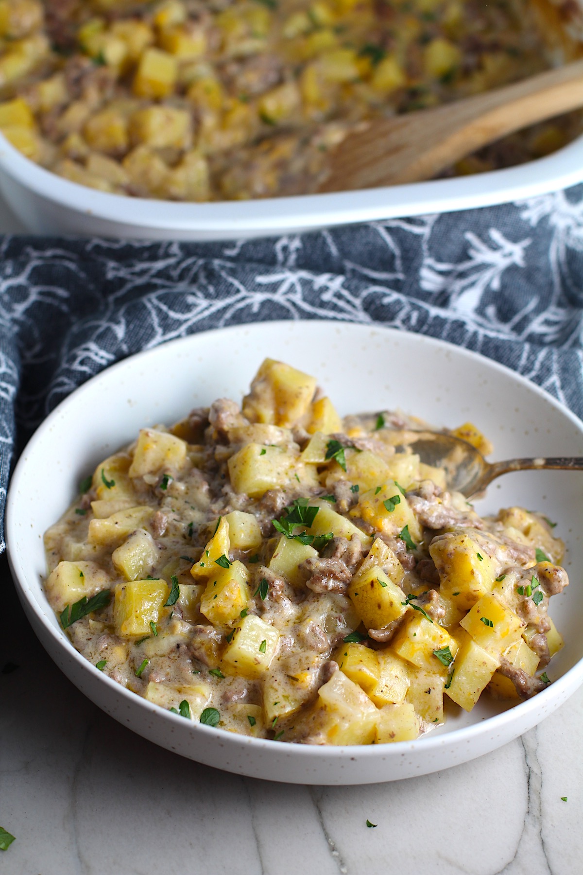 Photo o fHamburger Potato Casserole served in a white bowl with a spoon with a white baking dish with the casserole behind the bowl, by Talking Meals