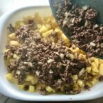 Pan pouring cooked ground beef and onions over diced raw potatoes for Cheesy Hamburger Potato Casserole. It's an easy, yummy, and cozy weeknight family dinner. 
