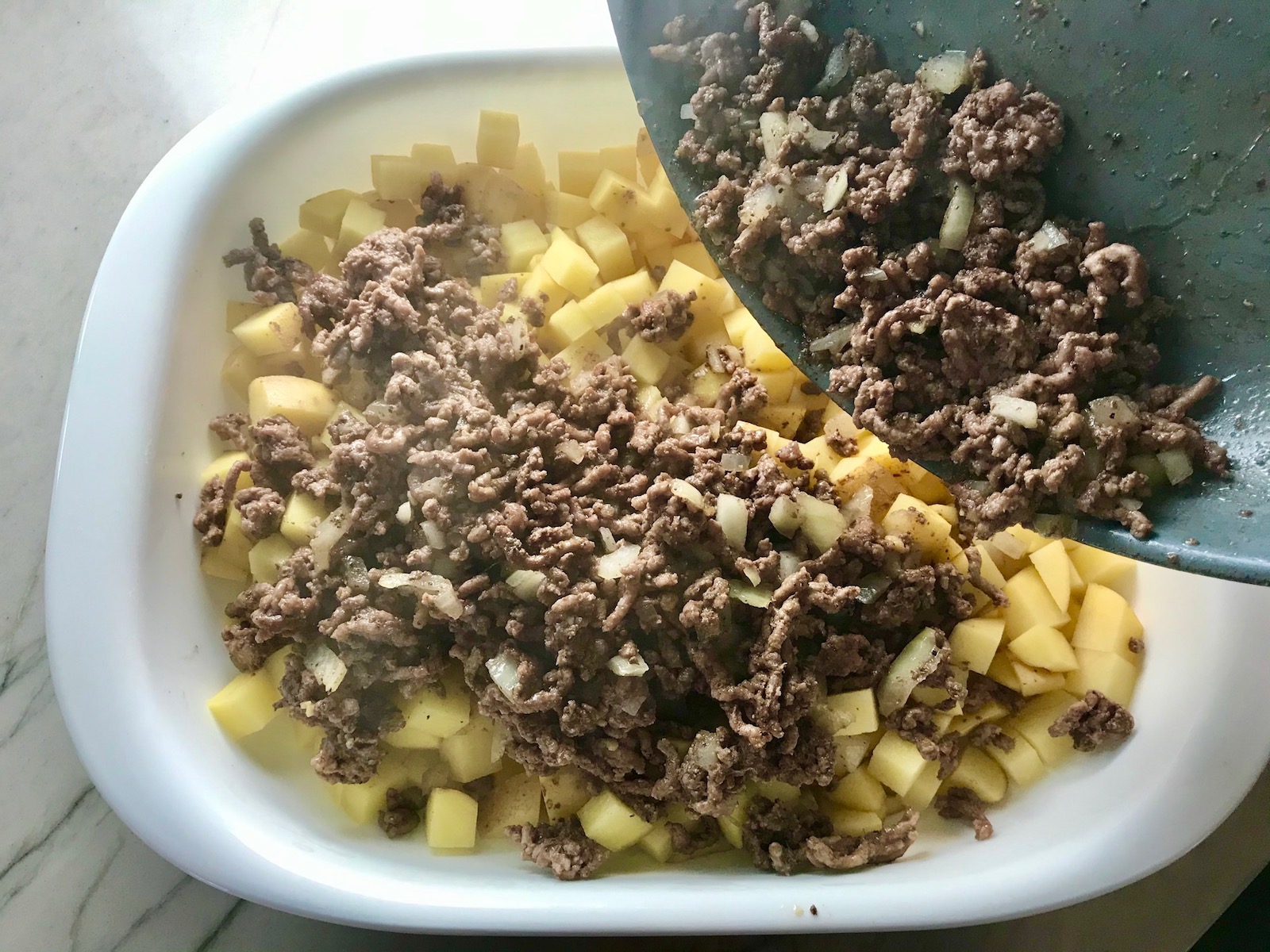 Pan pouring cooked ground beef and onions over diced raw potatoes for Cheesy Hamburger Potato Casserole. It's an easy, yummy, and cozy weeknight family dinner. 