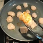 Tongs holding a scallop over pan seared side up for Glazed Pan Seared Scallops with Garlic and Honey.