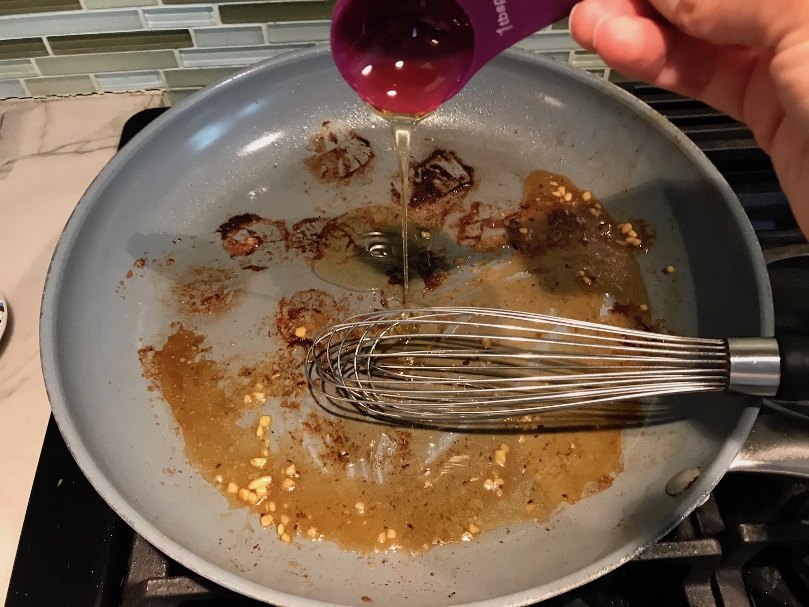 Hand pouring honey into pan with browned bits on bottom and garlic for Glazed Pan Seared Scallops with Garlic and Honey.