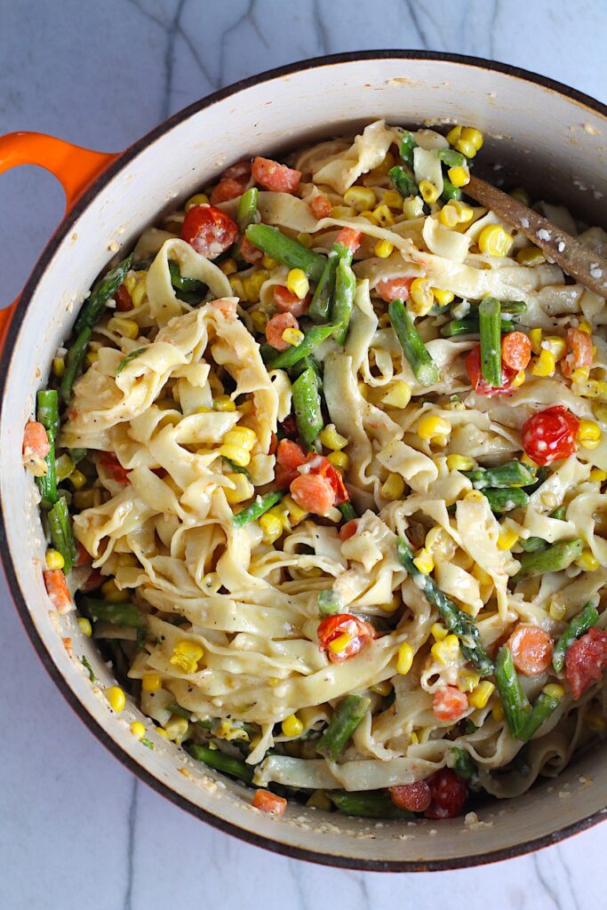 Fresh, Creamy Pasta Primavera Recipe in a pot on counter with fettuccine, parmesan cheese, roasted asparagus, carrots, tomatoes and fresh corn kernels.