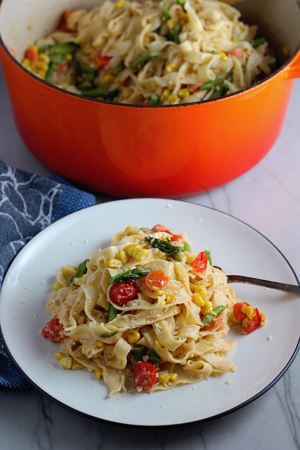 Fresh, Creamy Pasta Primavera Recipe on a plate with fork and pot in background on counter. It has fettuccine, parmesan cheese, roasted asparagus, carrots, tomatoes and fresh corn kernels.