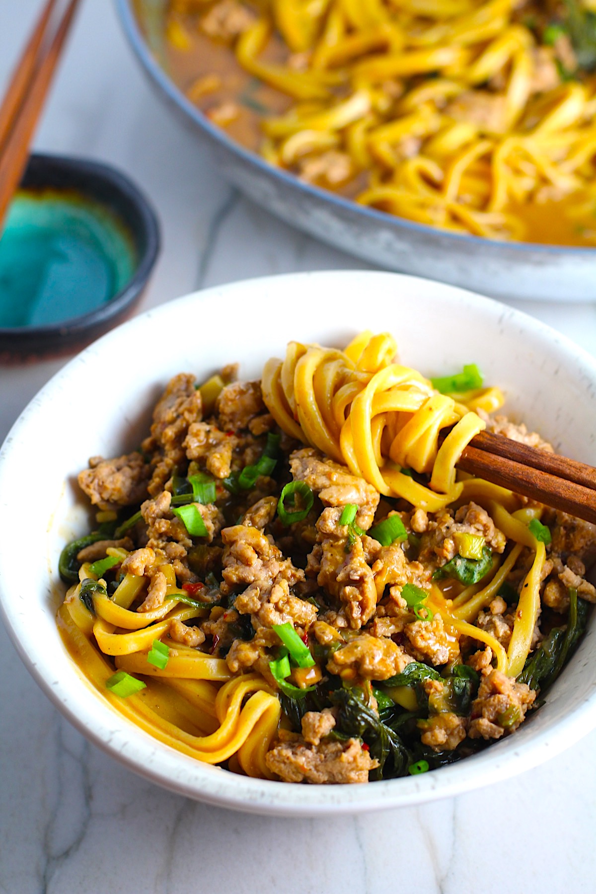 Dan Dan Noodle Recipe with Ground Chicken in a bowl. Chopsticks have noodles wrapped around and scallion slices are on top. The pan of noodles is in background.