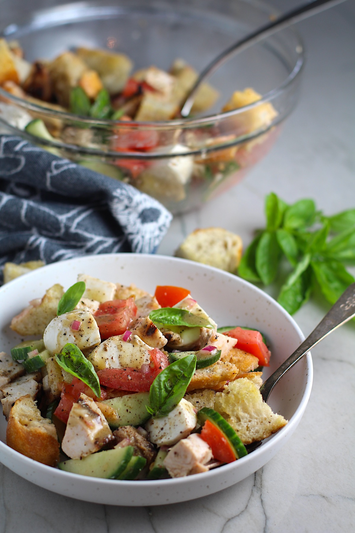Chicken Panzanella Salad Recipe in a bowl with fork.  It has Toasted bread cubes, chicken, tomatoes, cucumber, basil, and mozzarella.  Serving bowl in back
