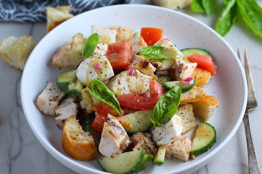 Toasted bread cubes, chicken, tomatoes, cucumber, basil, and mozzarella in a bowl for this Chicken Panzanella Salad Recipe. It's absolutely delicious.