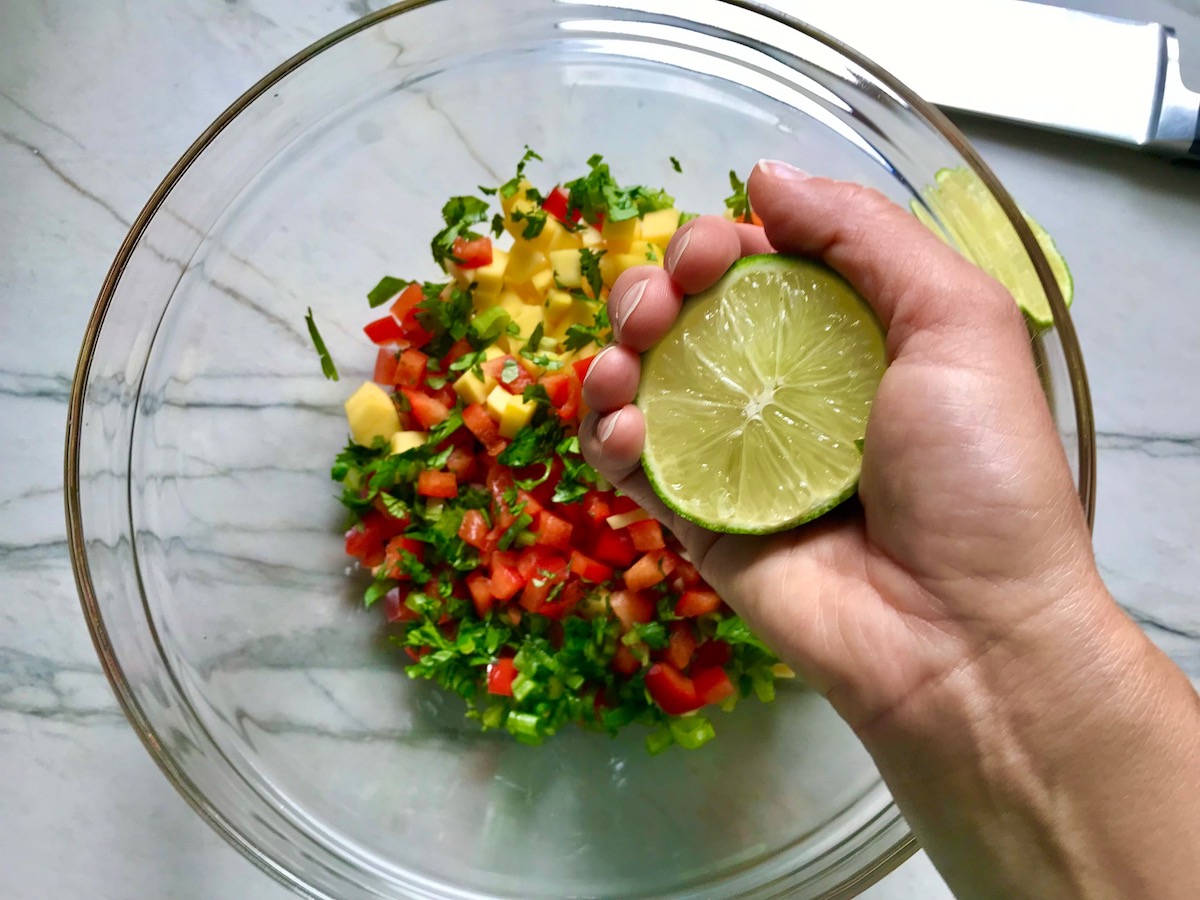Hand squeezing lime half over bowl of diced mango, red pepper, scallions, jalapeno, and cilantro for Grilled Shrimp Tacos