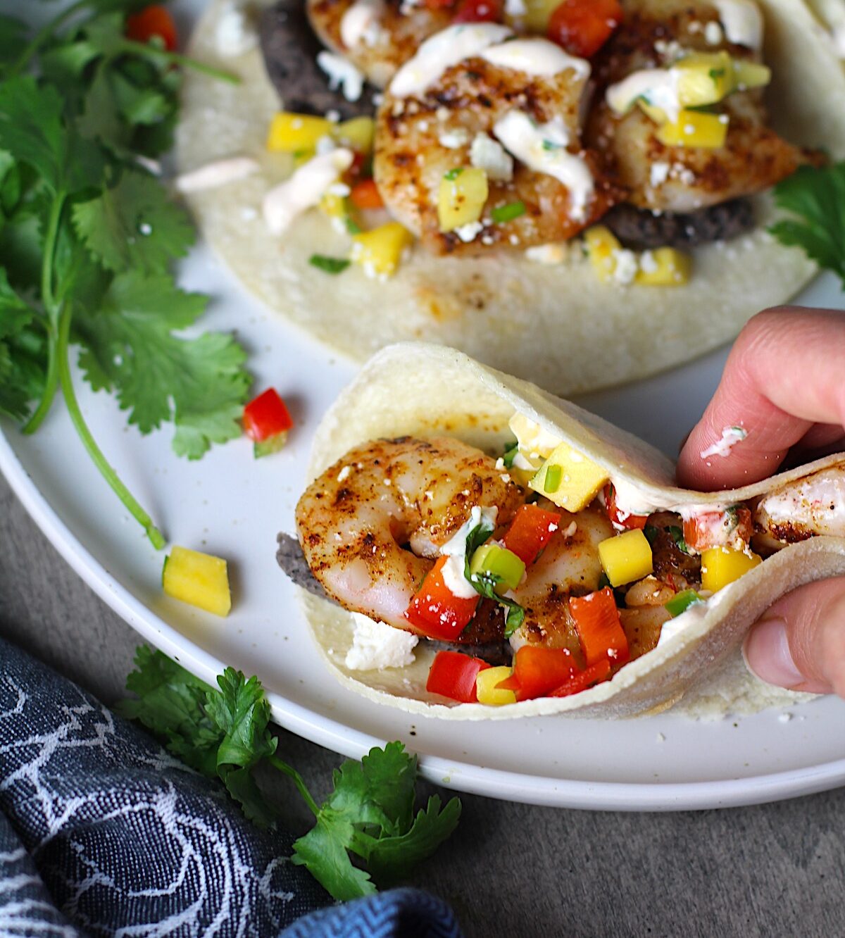 Hand holding a grilled Shrimp Taco in corn tortillas on a plate with Mango Salsa & Chipotle Crema. On the side are bowls of cotija cheese, crema, salsa, and cilantro leaves.