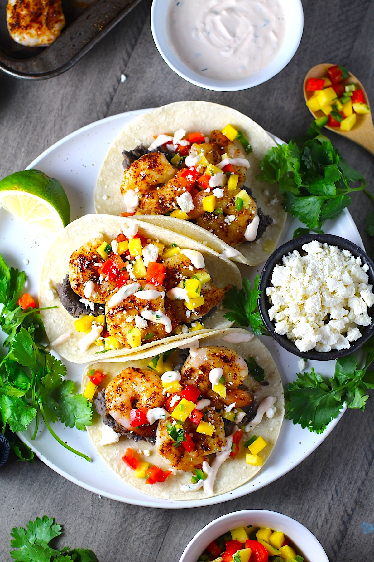 Grilled Shrimp Tacos in corn tortillas on a plate with Mango Salsa & Chipotle Crema.  On the side are bowls of cotija cheese, crema, salsa, and cilantro leaves. 