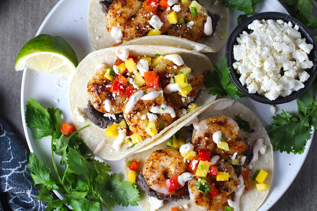 Grilled Shrimp Baja Tacos in corn tortillas on a plate with Mango Salsa & Chipotle Crema. On the side are bowls of cotija cheese, crema, salsa, and cilantro leaves.