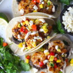 Grilled Shrimp Baja Tacos in corn tortillas on a plate with Mango Salsa & Chipotle Crema. On the side are bowls of cotija cheese, crema, salsa, and cilantro leaves.
