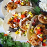 Grilled Shrimp Tacos in corn tortillas on a plate with Mango Salsa & Chipotle Crema. On the side are bowls of cotija cheese, crema, salsa, and cilantro leaves.