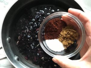 Hand holding small bowl with chili powder, cumin, and salt over pot with dried black beans for refried black beans recipe.