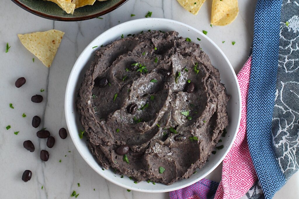 Creamy, rich, smokey, and delicious refried black beans recipe in a bowl with a few black beans and chopped cilantro sprinkled on top. Tortilla chips on the counter.