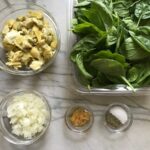 Prepped ingredients on counter for Spinach Artichoke Chicken Stuffed Pitas. From left top to right: chopped artichoke hearts, baby spinach leaves, diced onion, minced garlic, salt and pepper.