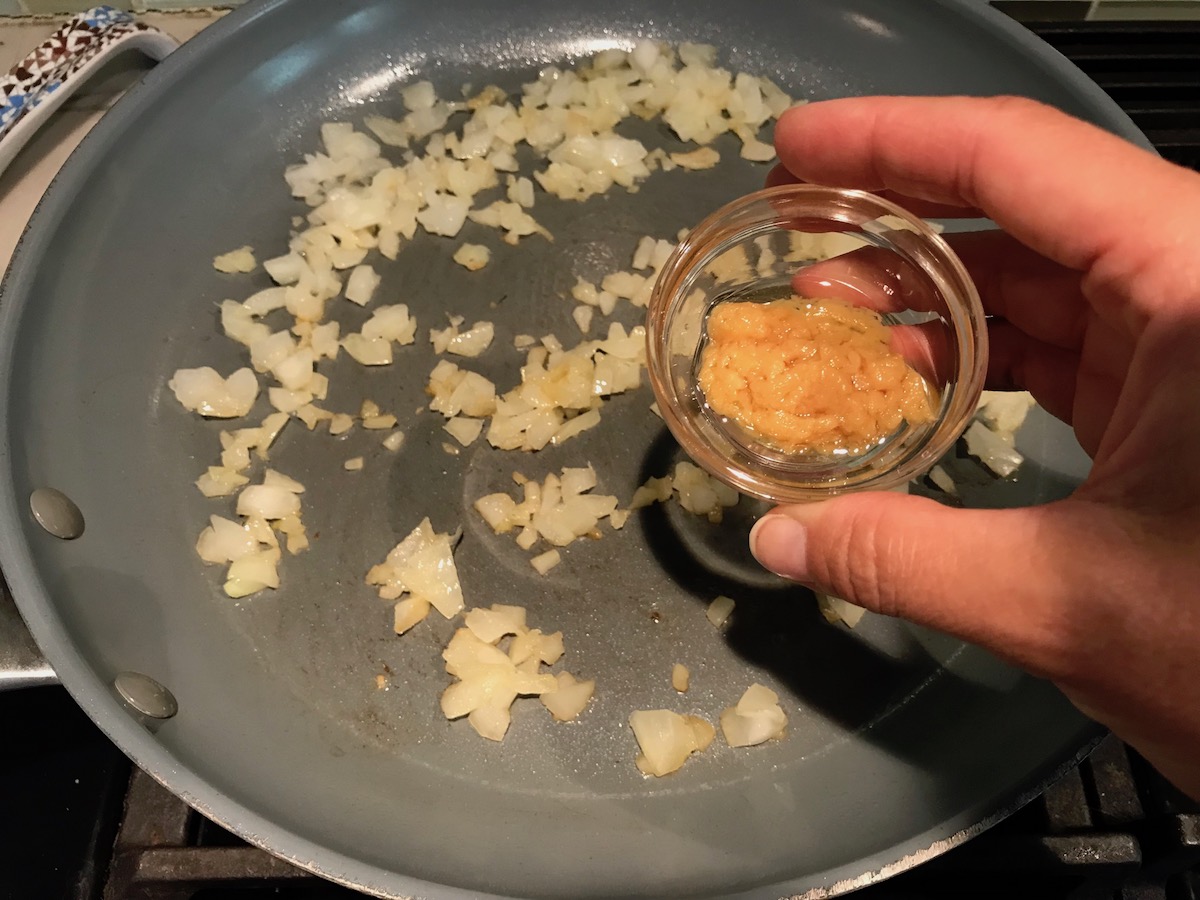 Hand adding minced garlic to cooking onions in a pan for the Spinach Artichoke Chicken filling that will be stuffed into pitas and then grilled.
