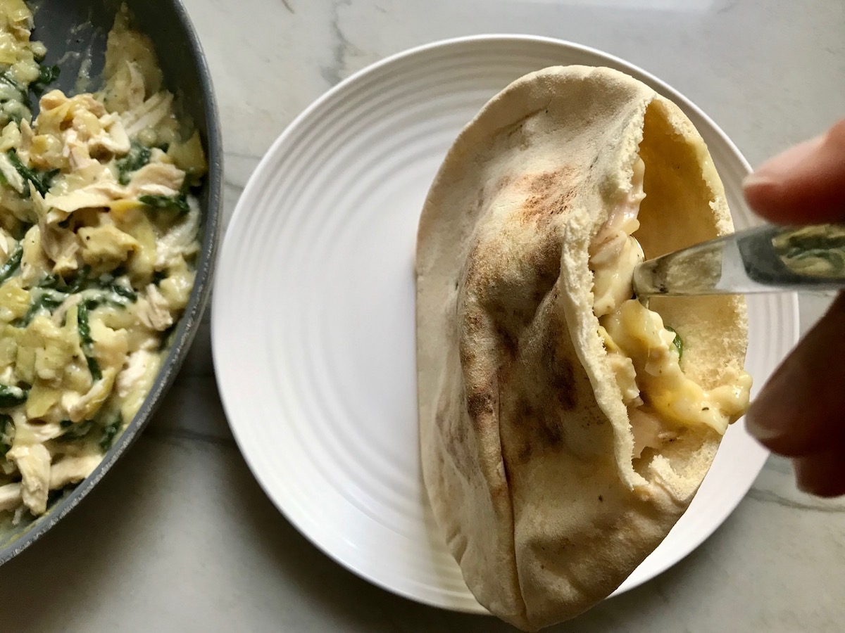 Spoon adding Spinach Artichoke Chicken filling to a cut open pita with pan of filling off to the side.