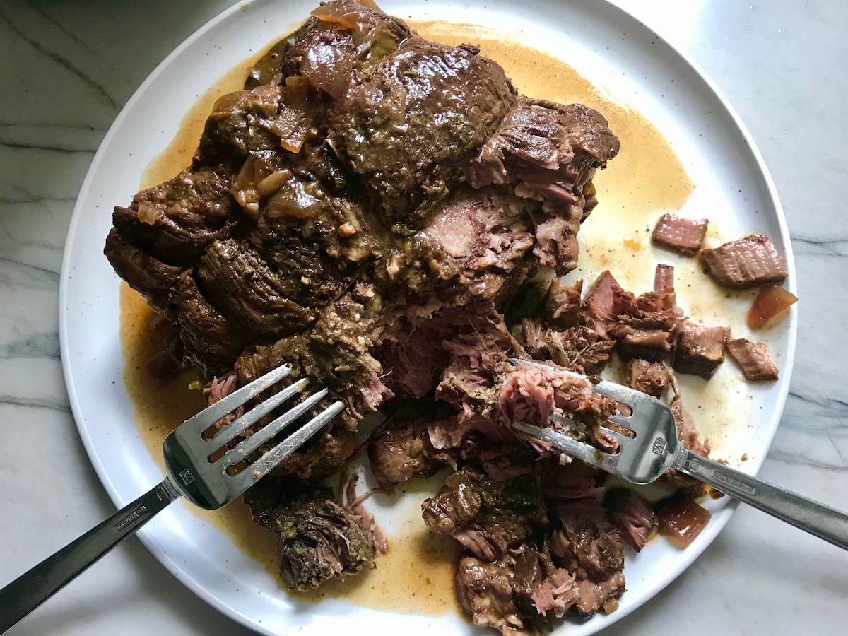 Two forks shredding beef chuck roast on a plate for Birria Tacos. This Birria Tacos Recipe in the Slow Cooker has shredded beef and Oaxaca cheese melted together in corn tortillas in a pan. 