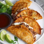 Four tacos fanned out on a plate for this Birria Tacos Recipe. Each taco has shredded beef and oaxaca cheese oozing out. On the plate is a bowl of red sauce, lime wedges, and cilantro.