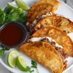 Four tacos fanned out on a plate for this Birria Tacos Recipe. Each taco has shredded beef and oaxaca cheese oozing out. On the plate is a bowl of red sauce, lime wedges, and cilantro.