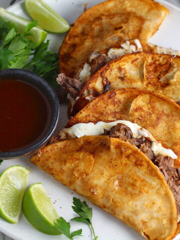 Four tacos fanned out on a plate for this Birria Tacos Recipe. Each taco has shredded beef and oaxaca cheese oozing out. On the plate is a bowl of red sauce, lime wedges, and cilantro.