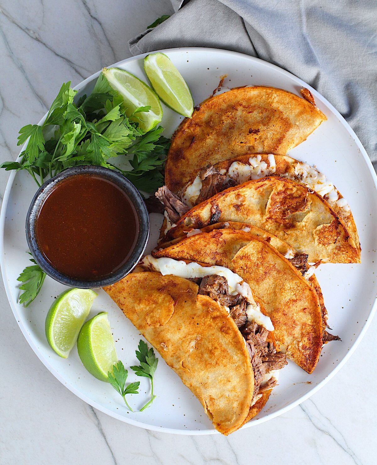 Four tacos fanned out on a plate for this Birria Tacos Recipe. Each taco has shredded beef and oaxaca cheese oozing out. On the plate is a bowl of red sauce, lime wedges, and cilantro.