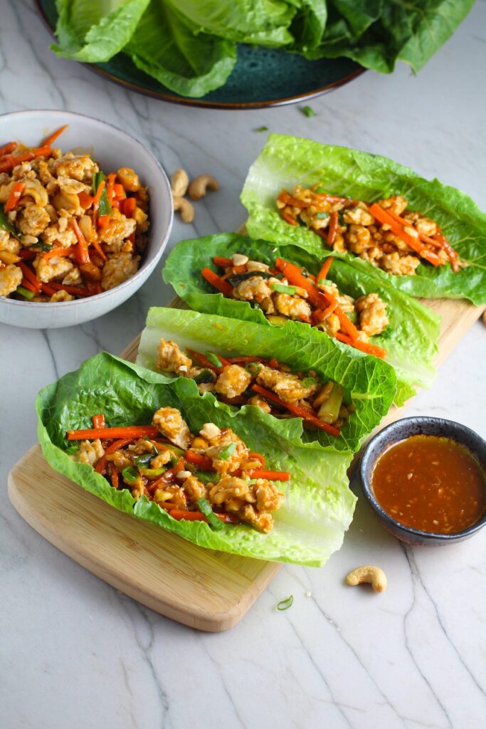 4 Cashew Chicken Lettuce Wraps on counter with a bowl of Cashew Chicken and Carrot filling on left and sauce on right.