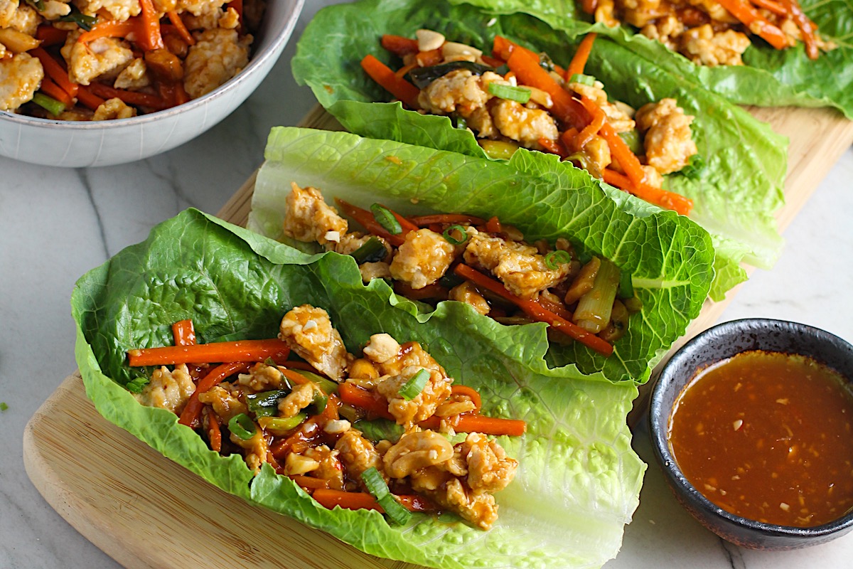 4 Cashew Low Carb Chicken Lettuce Wraps on counter with a bowl of Cashew Chicken and Carrot filling on left and sauce on right.