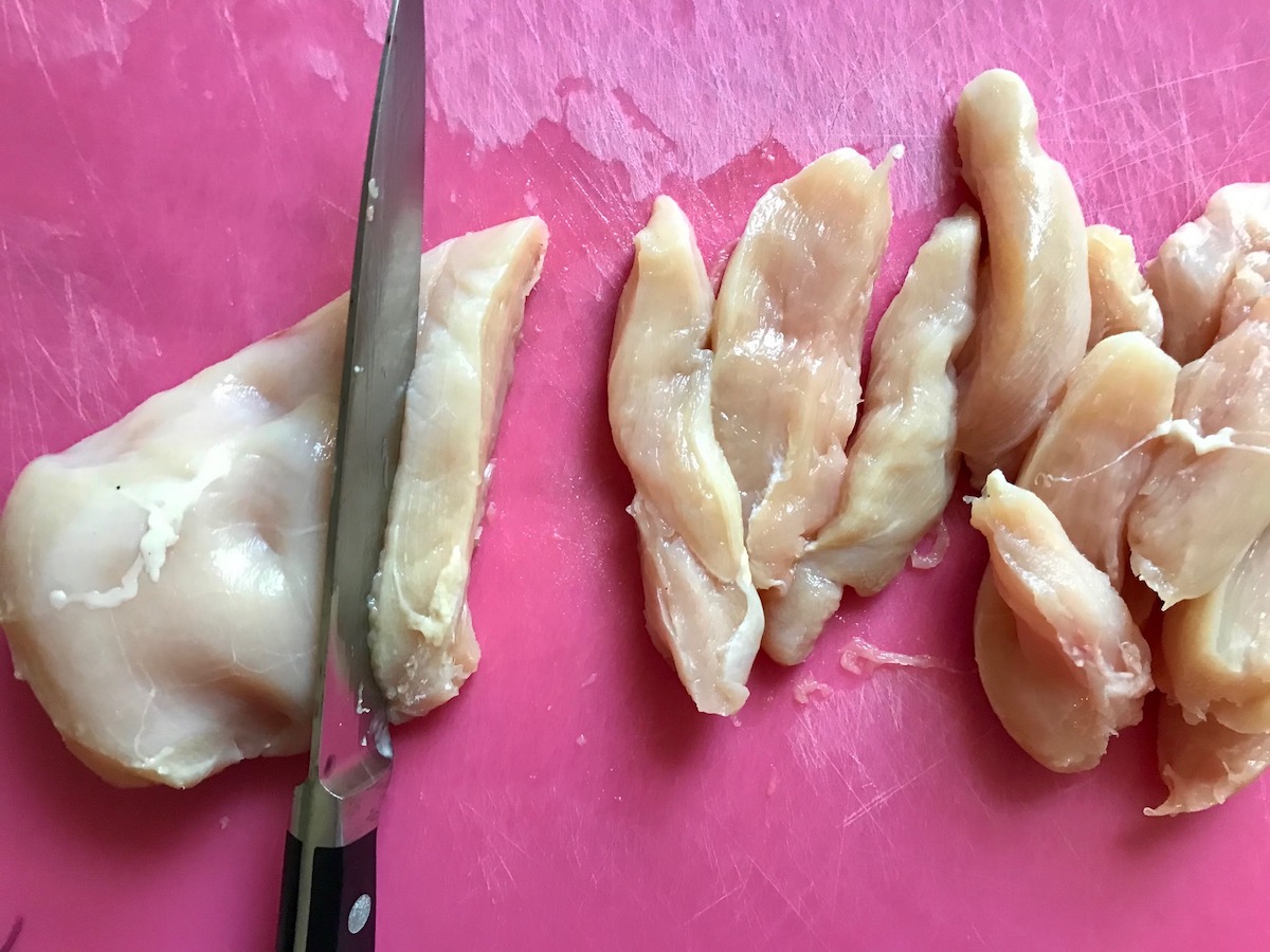 Knife cutting raw chicken breast into thin strips on a pink cutting board for Kale Caesar Salad with Baked Crispy Lemon Chicken strips