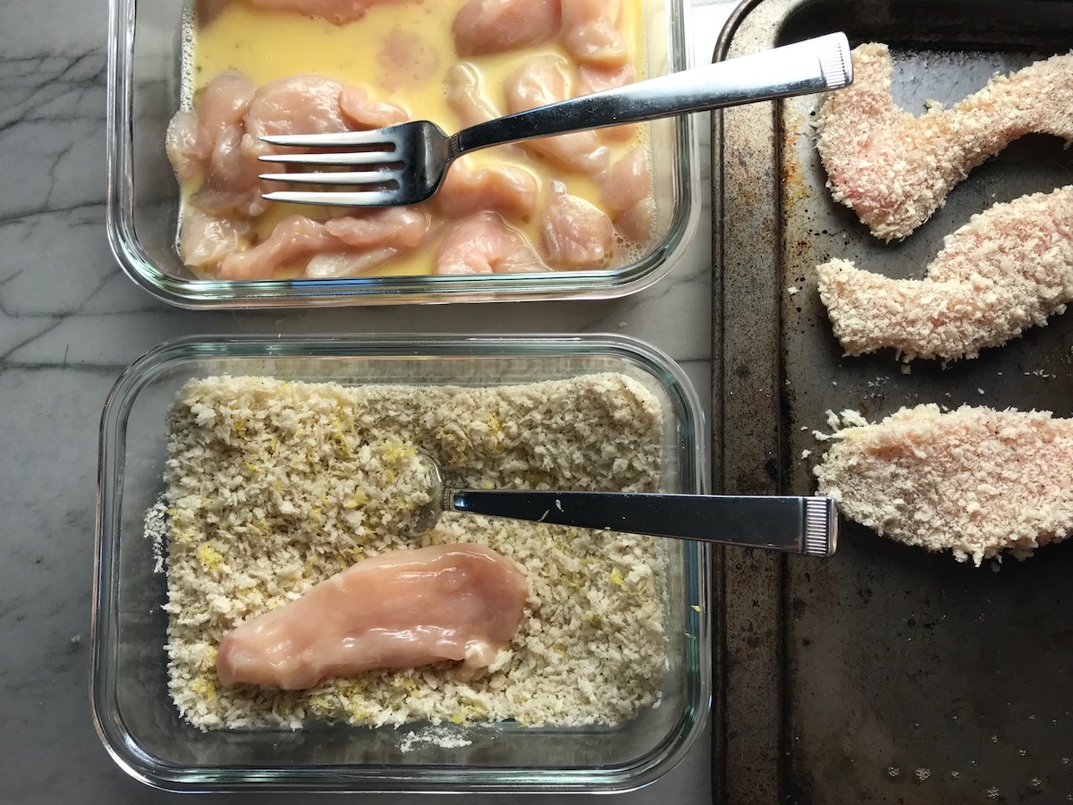 Dish with breadcrumbs and a raw chicken strip on left bottom, above that a dish with beaten eggs with raw chicken strips, and on the right is a sheet pan with breaded chicken strips for Kale Caesar Salad with Baked Crispy Lemon Chicken strips