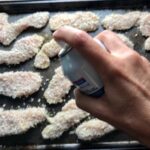 Hand spraying olive oil over breaded chicken strips on a sheet pan for Kale Caesar Salad with Baked Crispy Lemon Chicken strips