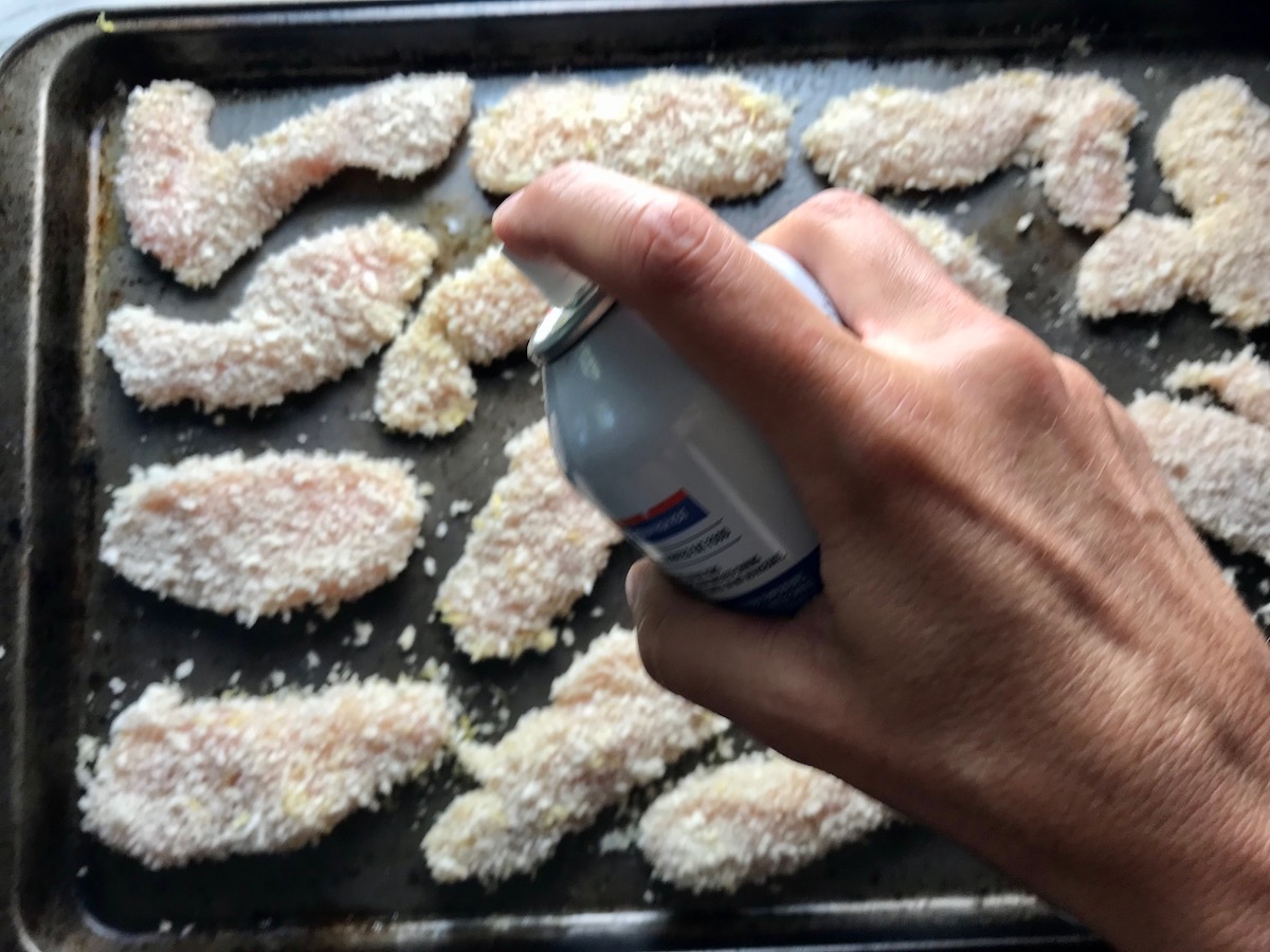 Hand spraying olive oil over breaded chicken strips on a sheet pan for Kale Caesar Salad with Baked Crispy Lemon Chicken strips
