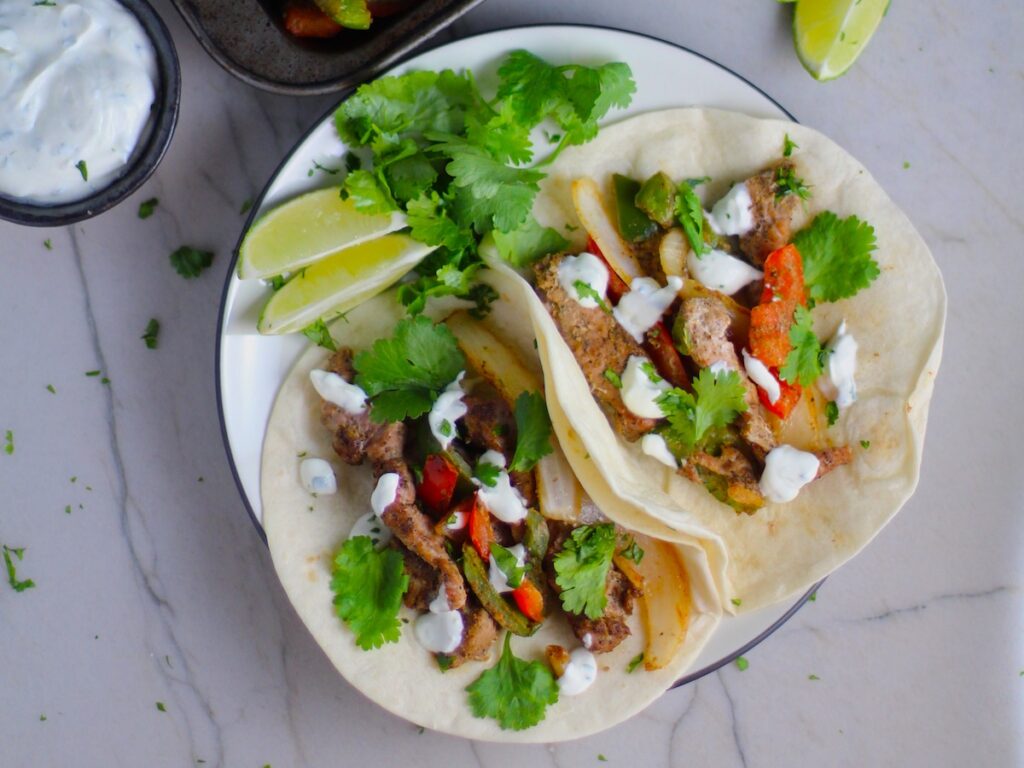 Sheet Pan Pork Fajitas Recipe with red pepper, green pepper, and onion, in flour tortillas on a plate. Topped with Cilantro Lime Crema, fresh cilantro leaves, and lime wedges.