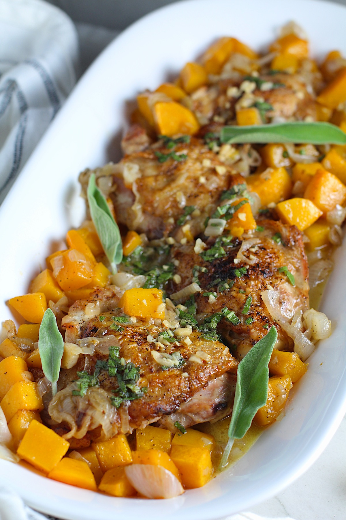 Butternut Squash and Chicken with sage, garlic, and shallots on a platter with fresh sage leaves and topped with a buttery garlic, lemon, sage sauce. Lemon wedges, fresh sage leaves, and a kitchen towel on counter.