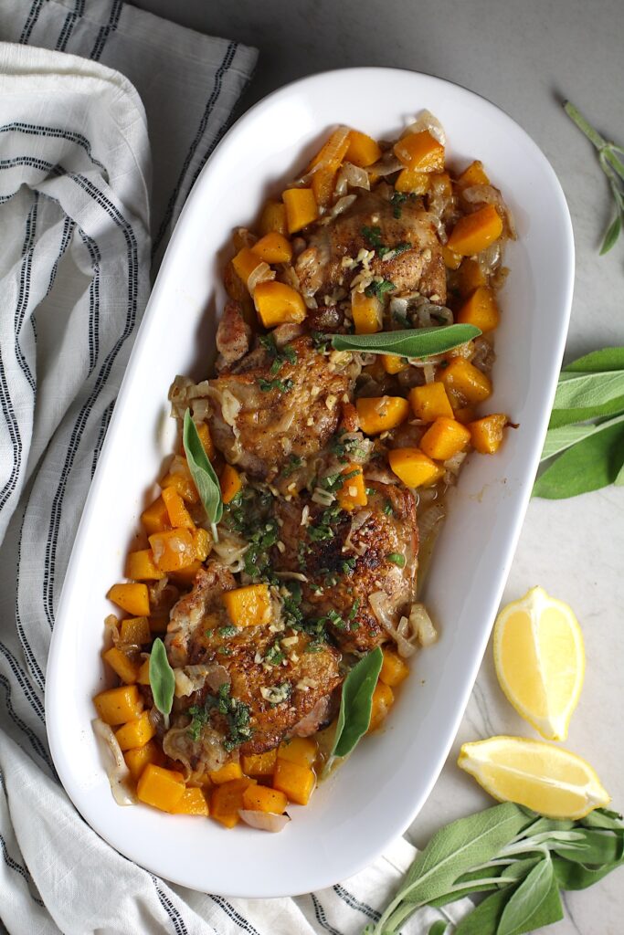 Crispy Sage Chicken, Butternut Squash, and Shallots on a platter with fresh sage leaves and topped with a buttery garlic, lemon, sage sauce. Lemon wedges, fresh sage leaves, and a kitchen towel on counter.