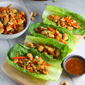 4 Low Carb Chicken Lettuce Wraps on counter with a bowl of Cashew Chicken and Carrot filling on left and sauce on right.