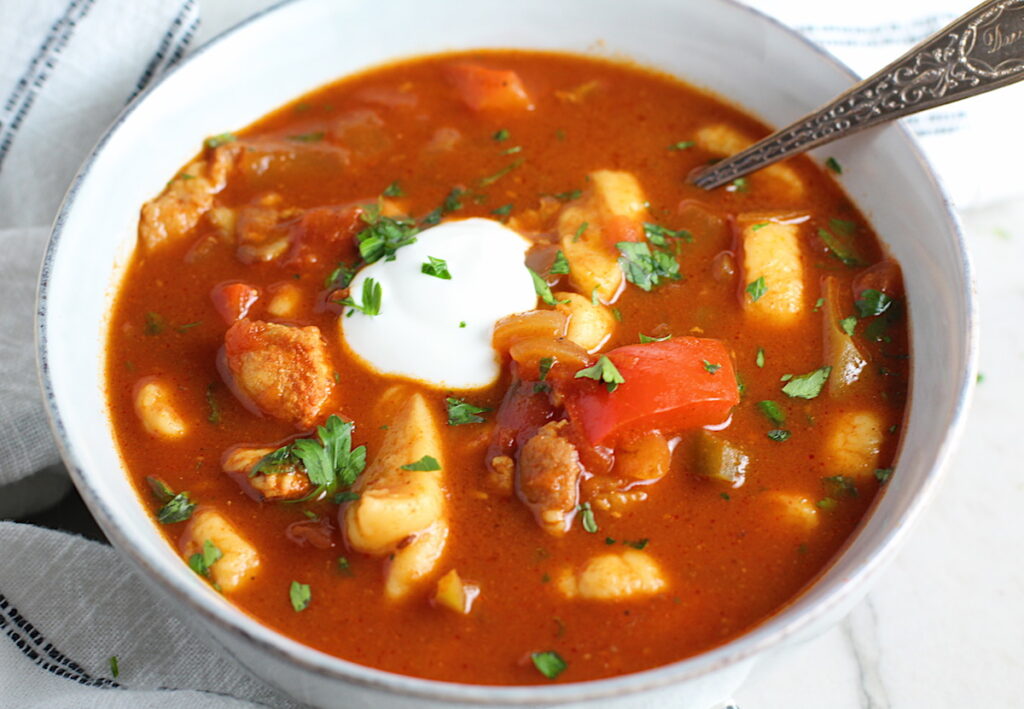 Chicken Goulash with quick homemade dumplings, red pepper, and parsley in a bowl with a spoon and sour cream on top.