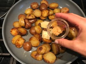 Hand adding minced garlic to skillet with 2 pats of butter and browned and crispy halved red and gold creamer potatoes for Sage Butter Potatoes.