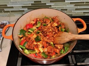 Wood spoon stirring red and green peppers, chicken, bacon and diced onions in pot cooking for Chicken Goulash with quick homemade dumplings.