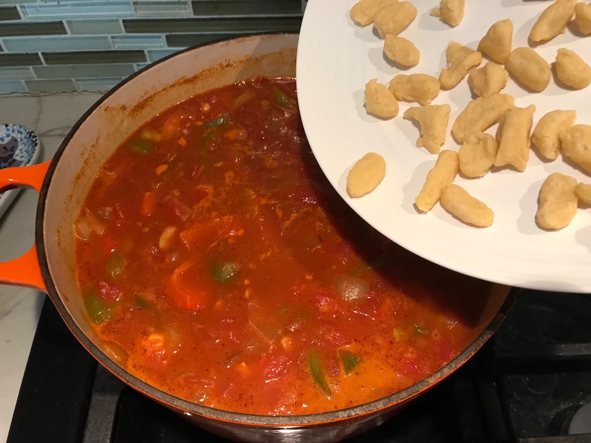 Uncooked dumplings on a plate being added to Chicken Goulash cooking in a pot on stove.