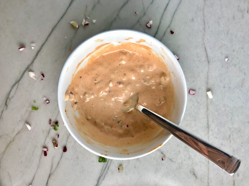 Pink-colored remoulade sauce in a bowl with a spoon on counter with chopped ingredients on the counter to go with Crispy Baked Cod Panko Fish and Chips.