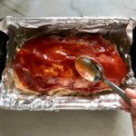 Spoon spreading ketchup over the top of raw Ground Chicken Meatloaf in a loaf pan that's lined with aluminum foil. It's a perfect family dinner main dish!  10 minutes to prep and 45 minutes to bake.