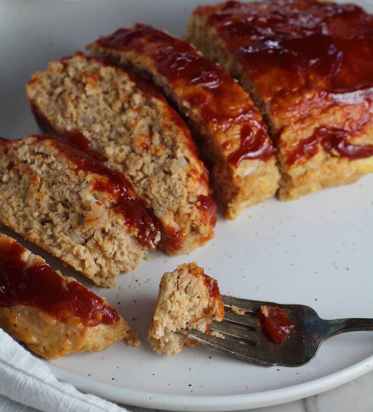 Ground Chicken Meatloaf with ketchup on top sliced and fanned out on plate with fork and a piece on side and towel next plate. It's a perfect family dinner main dish!  10 minutes to prep and 45 minutes to bake.