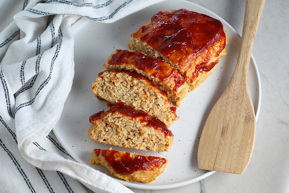 Ground Chicken Meatloaf with ketchup on top sliced and fanned out on plate with spatula on side and towel next plate. It's a perfect family dinner main dish!  10 minutes to prep and 45 minutes to bake.