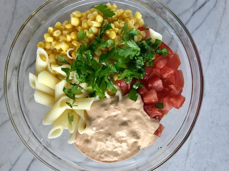 Clear bowl on counter with corn, tomatoes, pasta, Mexican dressing, and cilantro. This Creamy Mexican Pasta Salad has Sun Dried Tomatoes, Corn, Fresh Diced Tomatoes, Cilantro, and smoky Mexican spices. The dressing is creamy and smoky with lots of depth and a hint of sweetness from the Sun Dried Tomatoes that are blended in. Terrific as a side or mix in some chicken or beef and call it a meal!