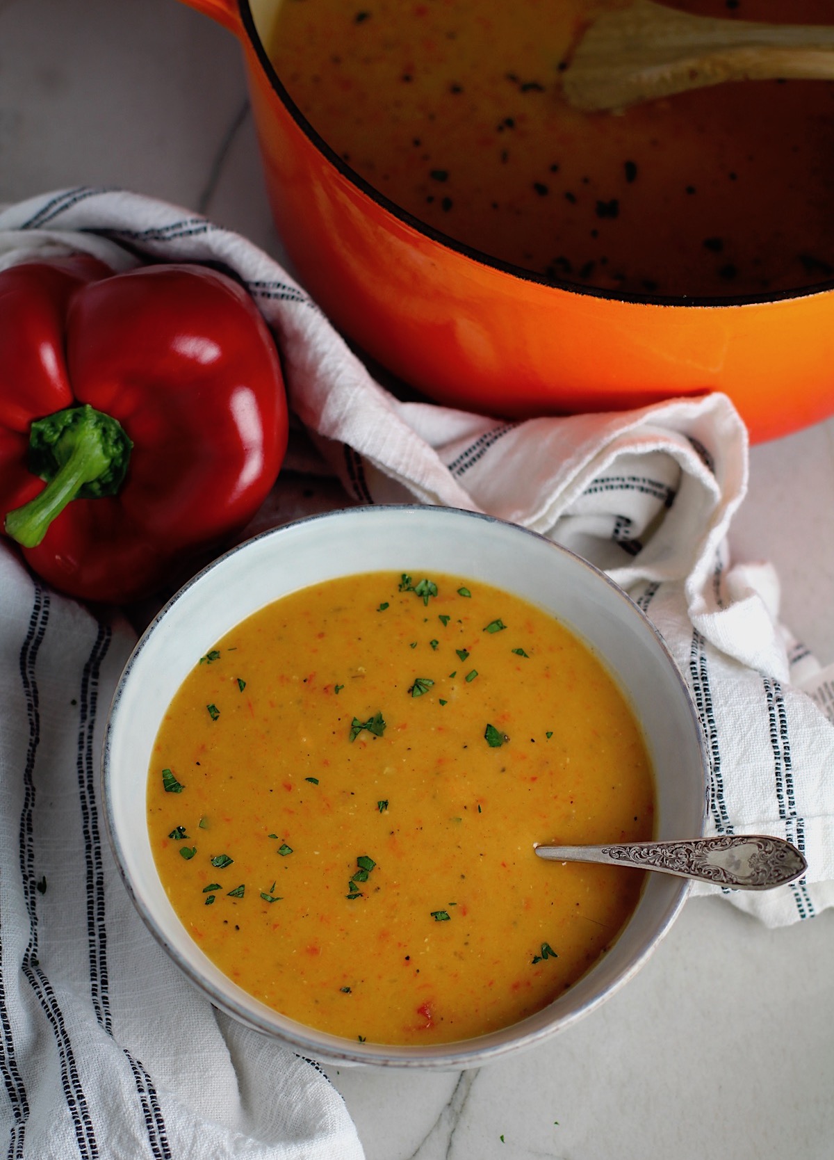 Creamy Red Pepper and Lentil Soup in a bowl with spoon and pot in background. Whole red pepper next to bowl. This is an easy and hearty lunch or dinner!  It comes together in just 40 minutes.
