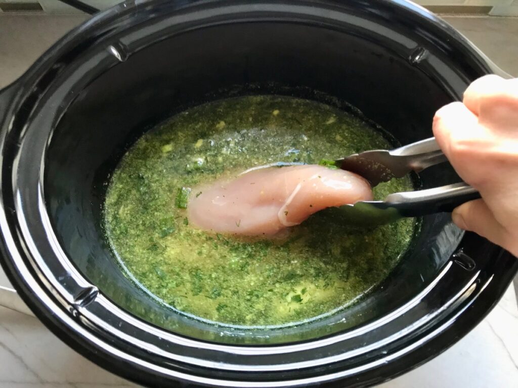 Tongs adding raw chicken breast to broth and herbs in crockpot for Ranch Chicken Crock Pot Pasta recipe. It uses fresh dill, parsley, and scallions is creamy, tangy, and utterly delicious!  It's also so easy to make, so it's a great weeknight dinner.