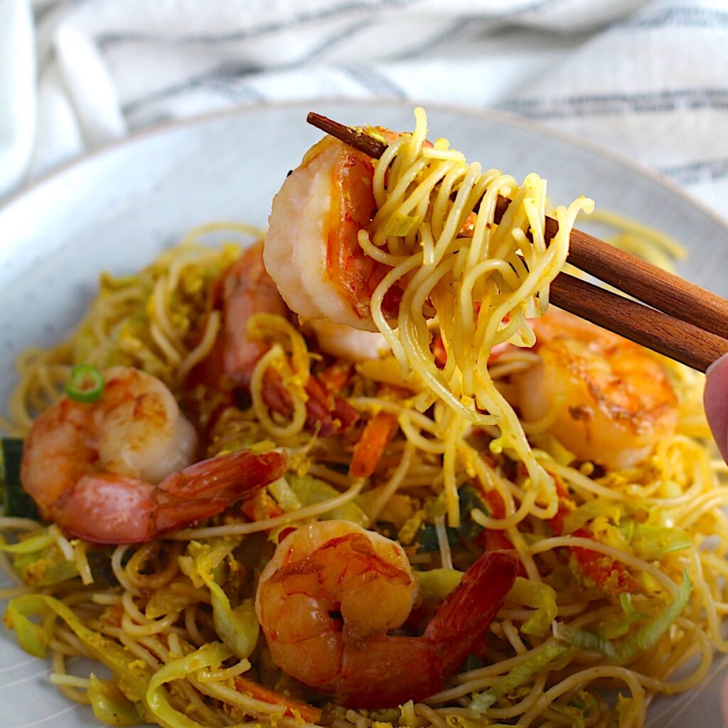 Chopsticks holding a bite of Shrimp Mei Fun Recipe, Singapore Noodles, over a plate on counter with napkin. It's an easy stir fry to make at home and tastes so much better than take out! With carrots, bean sprouts, curry powder, sesame oil, and more it's is a 30 minute weeknight dinner.